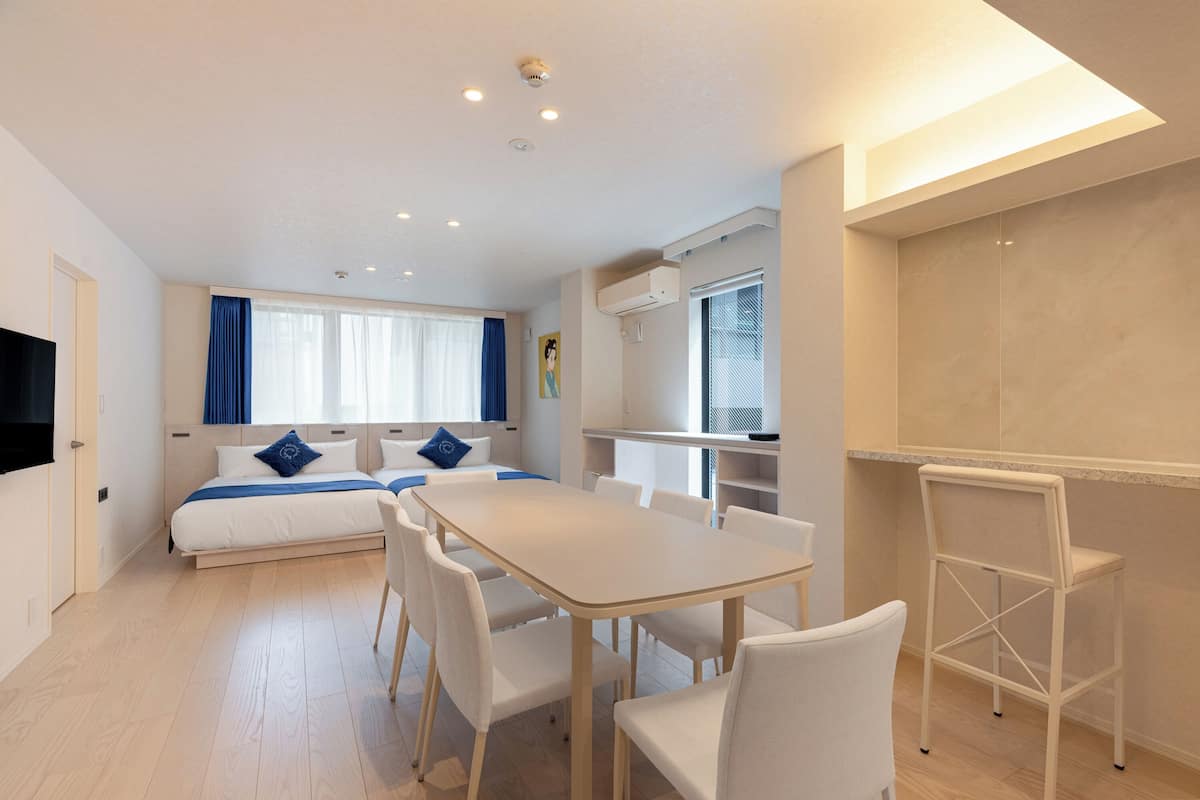 A modern hotel room with a bed, a dining table, chairs, a TV, and a kitchenette.
