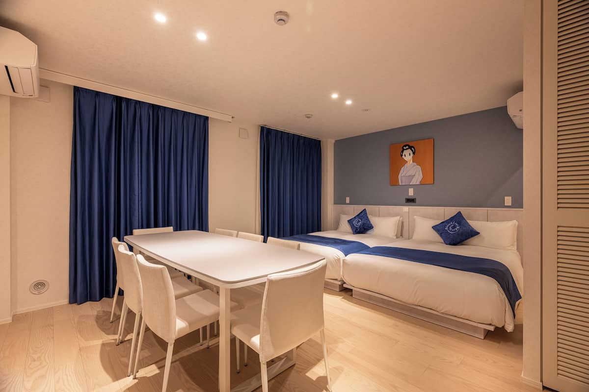 A modern hotel room with a dining table, chairs, a bed with blue bedding, and a painting on the wall.
