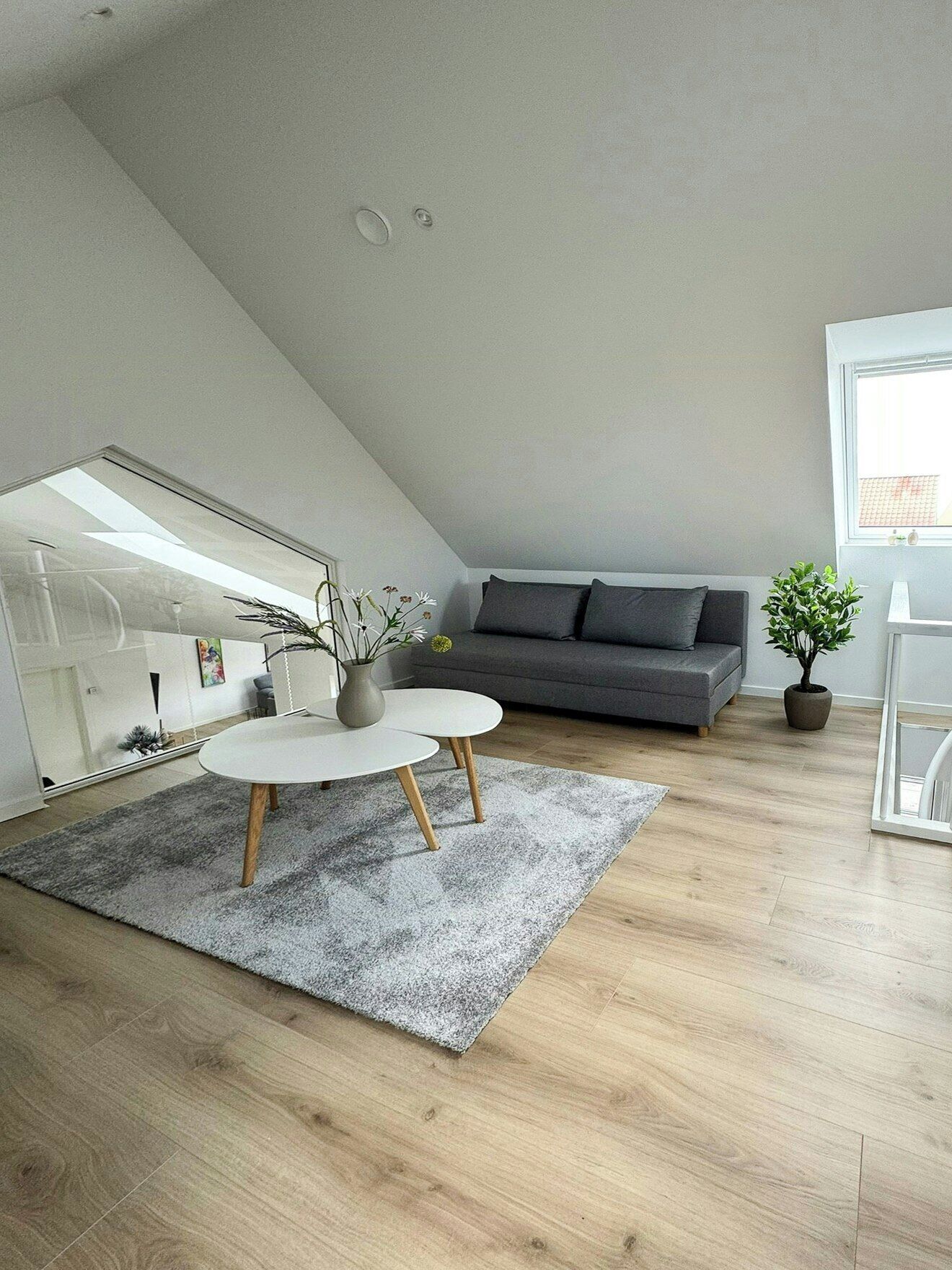 A modern loft bedroom with a sloped ceiling, a large window, a gray sofa, a round coffee table, and a potted plant.