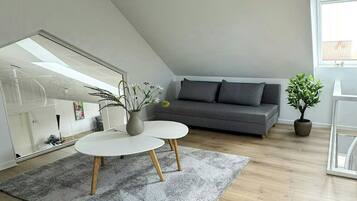 A modern loft bedroom with a sloped ceiling, a large window, a gray sofa, a round coffee table, and a potted plant.