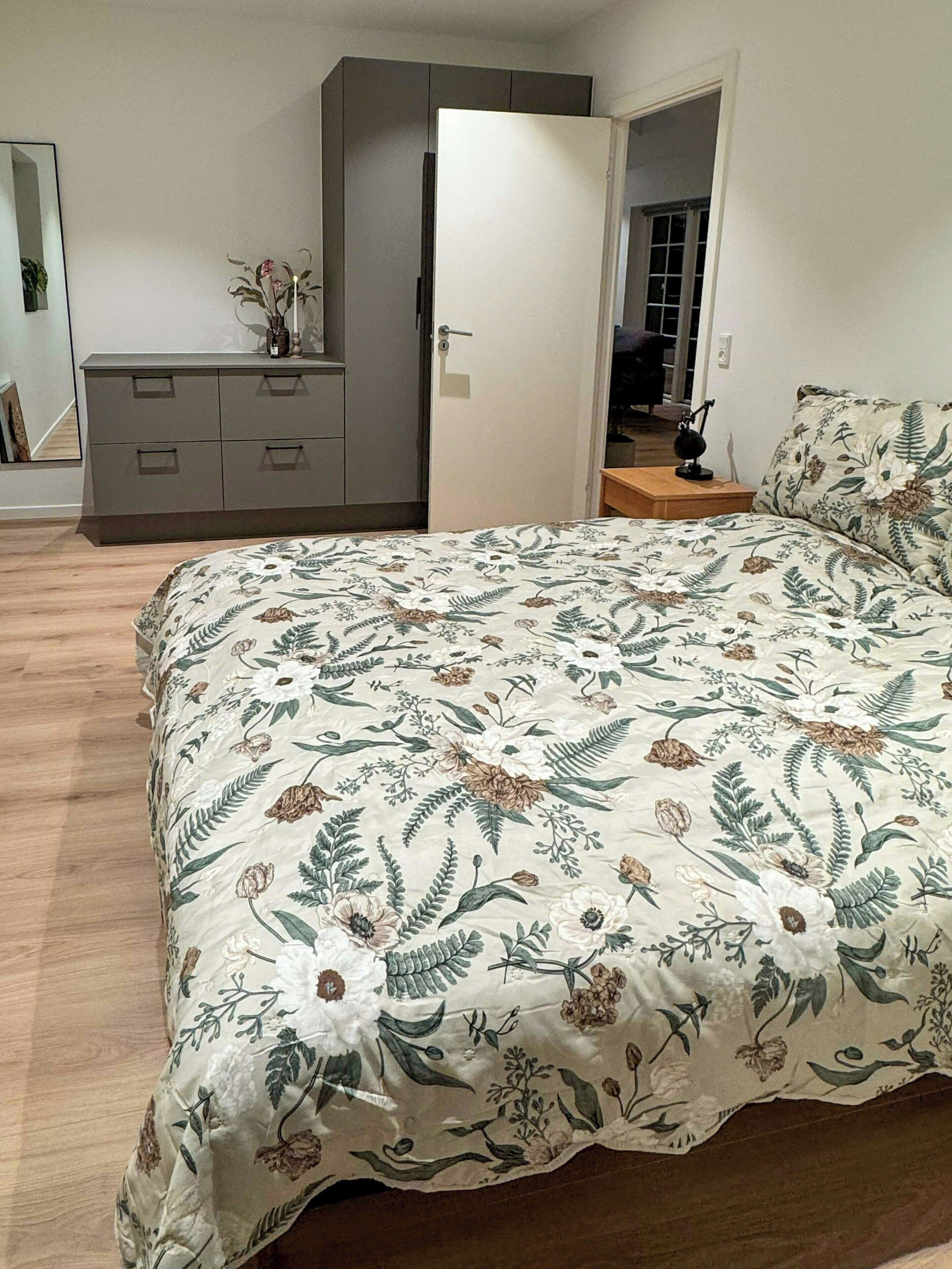 A bedroom with a floral bedspread, a grey dresser, and a door leading to another room.
