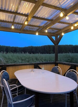Un patio al aire libre con una mesa redonda y sillas, luces colgantes y vistas al bosque.