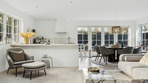 Une cuisine moderne avec des meubles blancs, un coin repas avec une table ronde et des chaises, et un salon avec un canapé et un pouf.