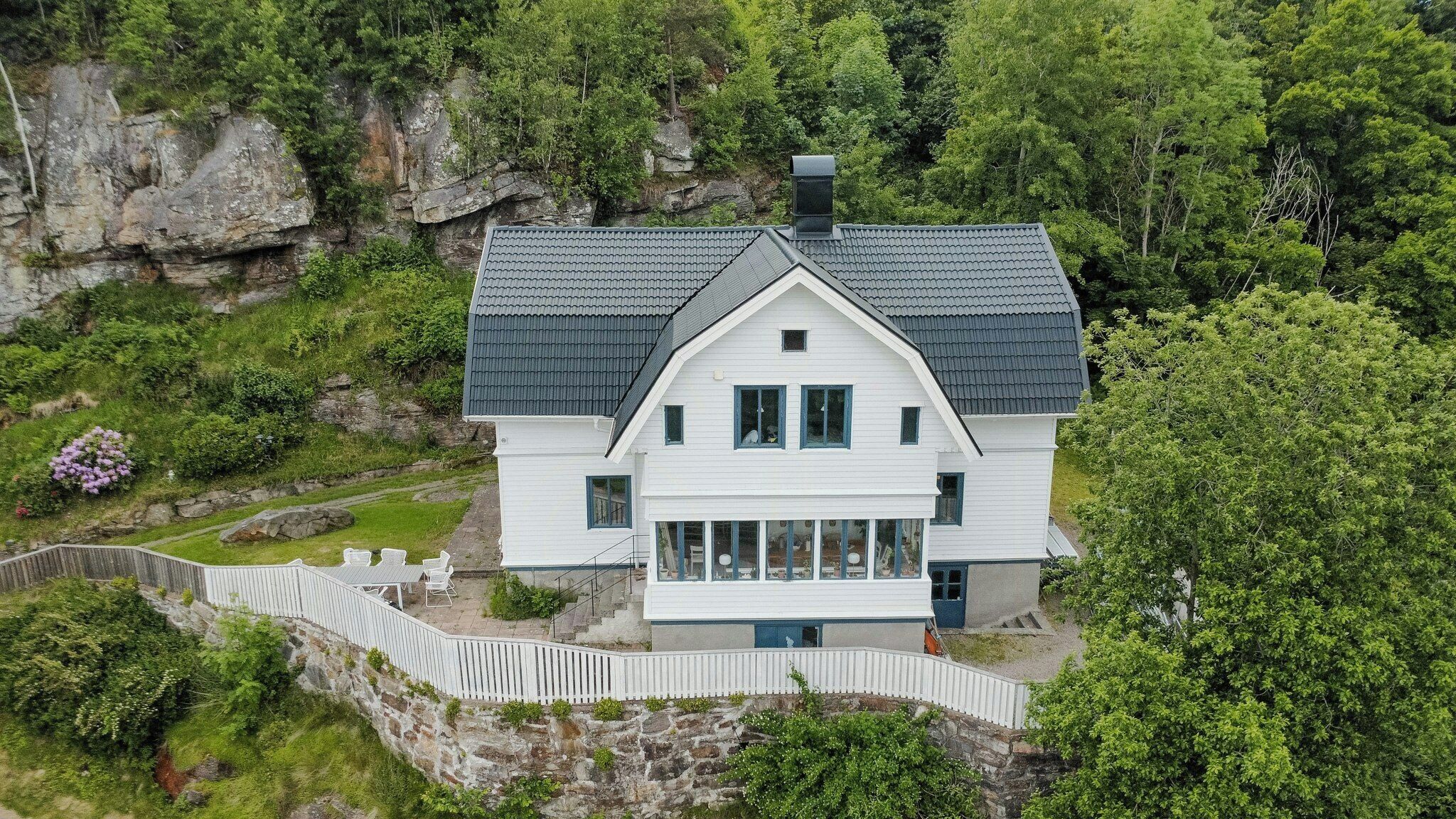 A white two-story house with a dark roof, surrounded by greenery and a stone wall.