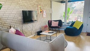 A living room with a brick wall, a flat-screen TV, a sofa, a blue chair, and a window with plants. - Colorful character and charm by Nakskov Fjord (Nakskov)