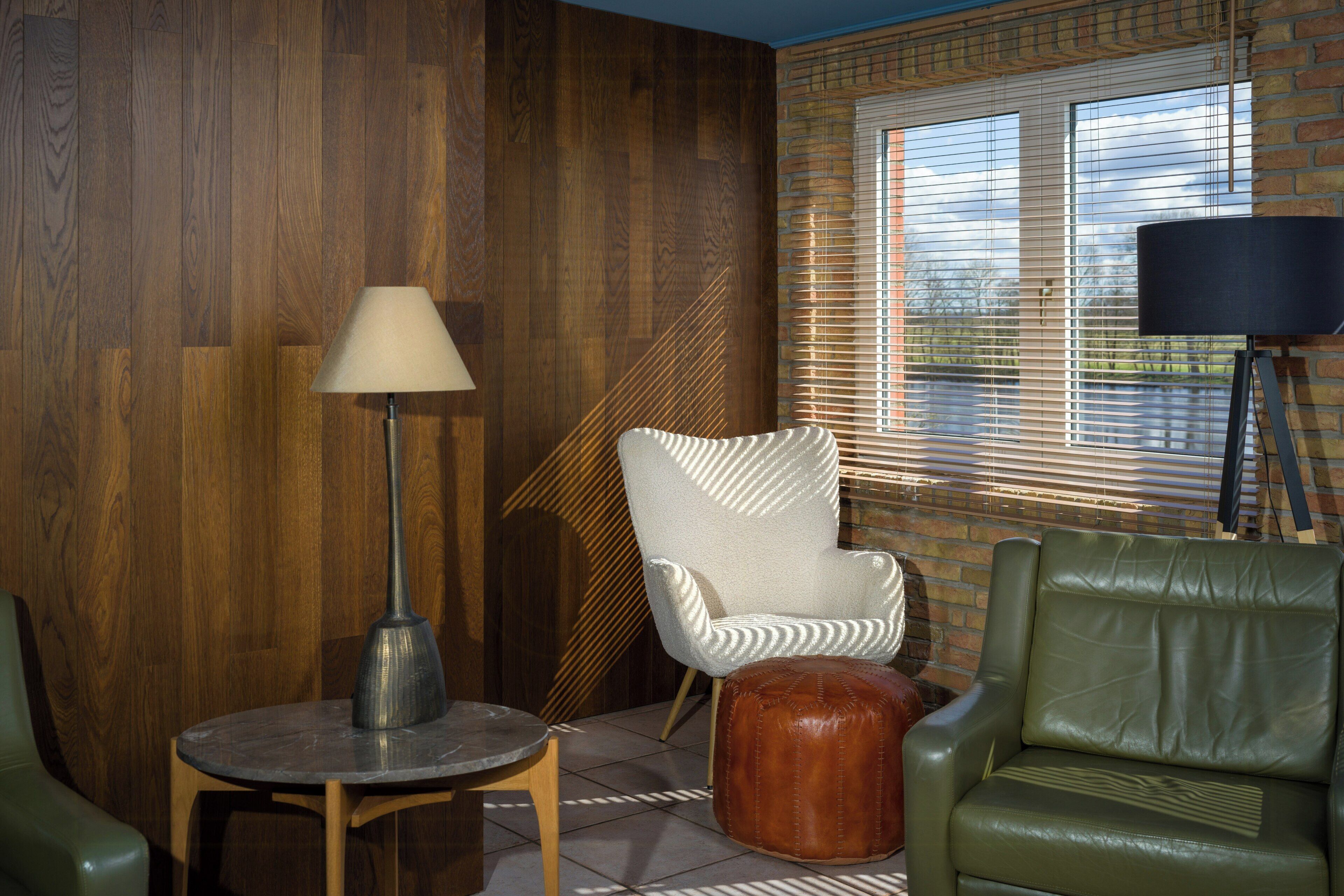 A room with a wooden wall, a round table with a lamp, a green leather chair, a white armchair, and a window with blinds.