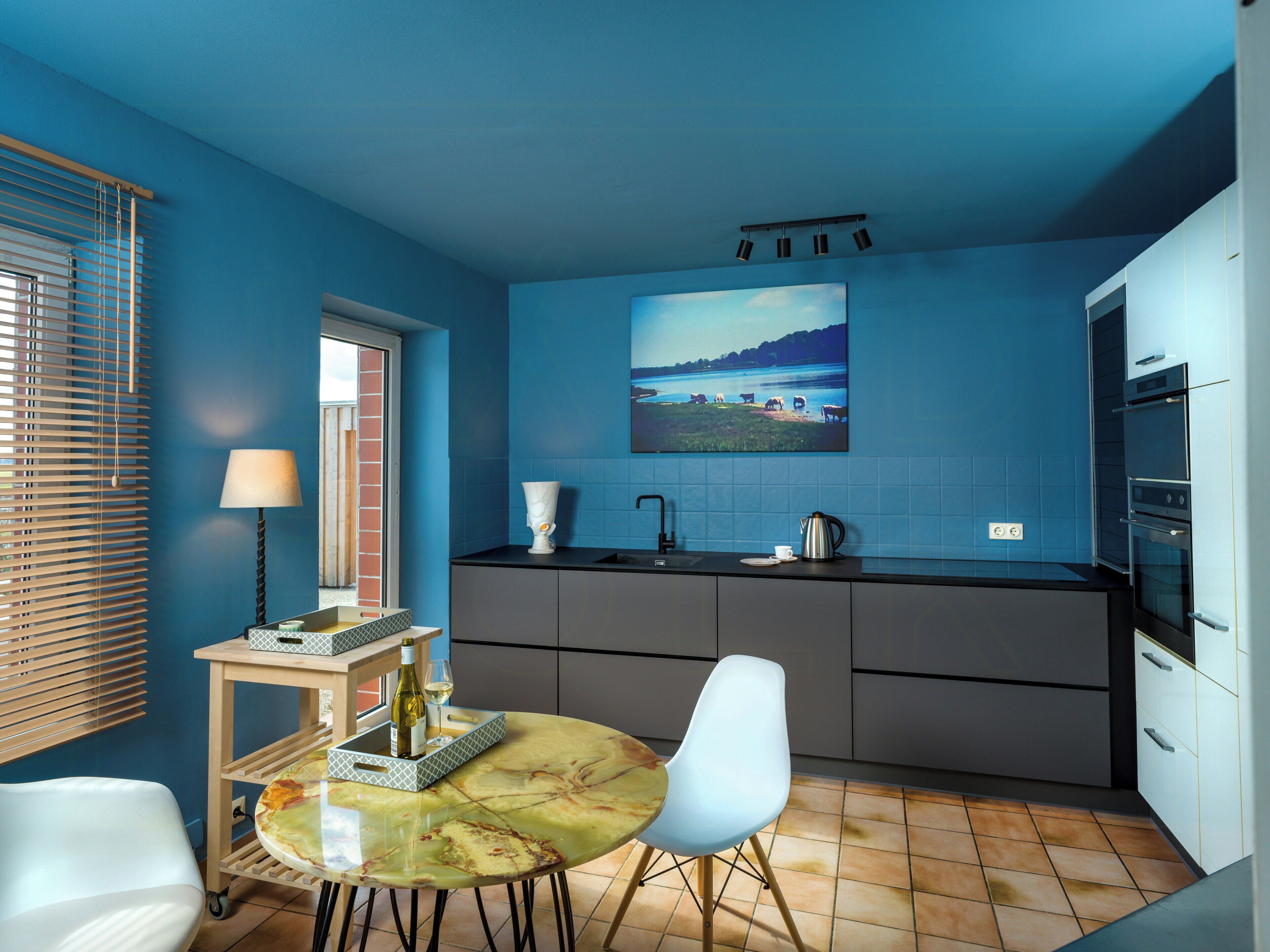A modern kitchen with a blue wall, a dining area with a table and chairs, and a window with blinds.