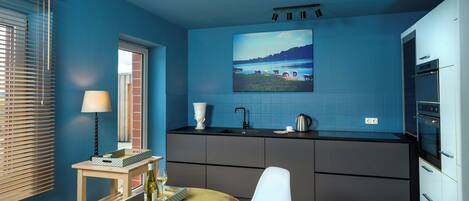A modern kitchen with a blue wall, a dining area with a table and chairs, and a window with blinds.