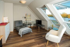 Un dormitorio tipo loft moderno con techo inclinado, un tragaluz grande, un sofá gris, un sillón blanco, una mesita con una botella de vino y un televisor sobre un mueble.