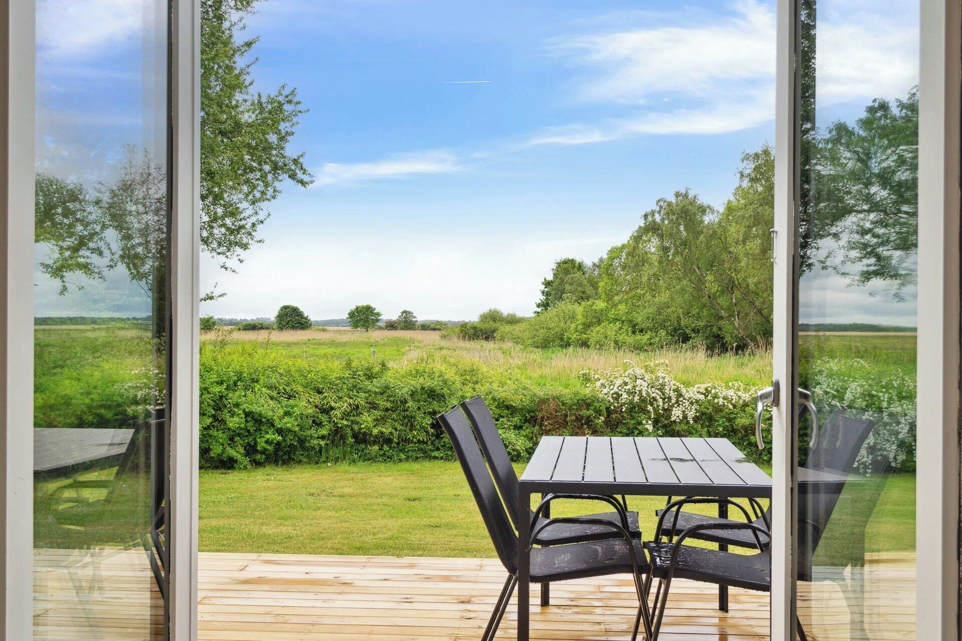 En træterrasse med et bord og stole, med udsigt over en græsplæne med træer og en klar himmel.