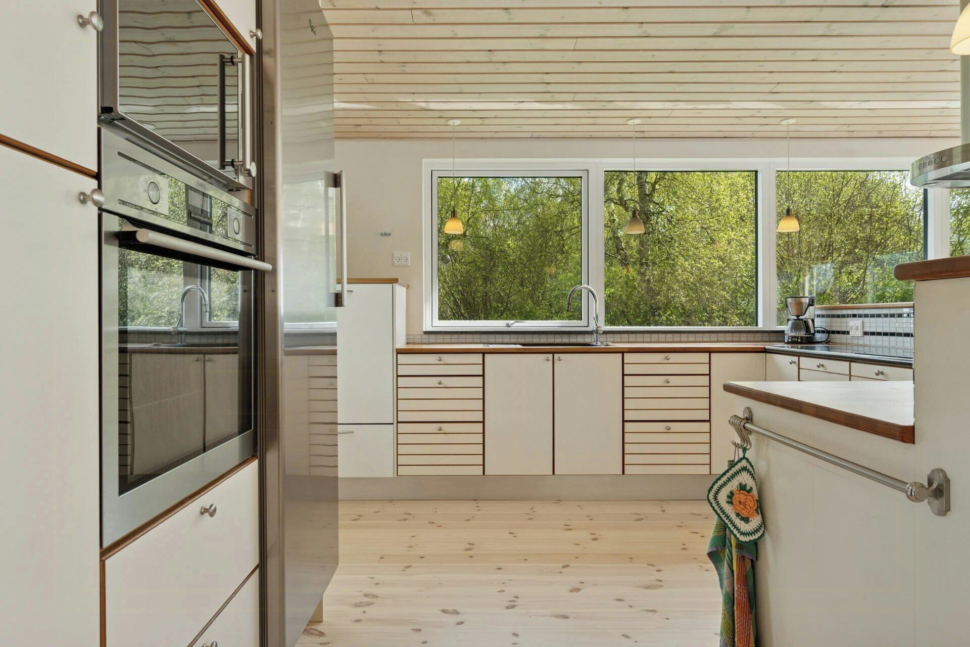 A modern kitchen with wooden cabinets, a built-in oven, a large window with a view of trees, and a hanging towel.