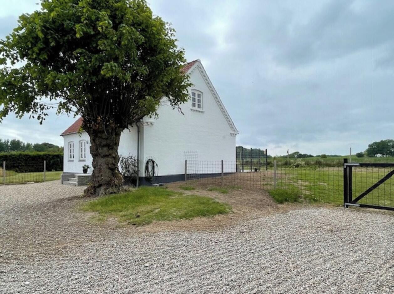 A white building with a large tree in front, surrounded by gravel and a metal fence.