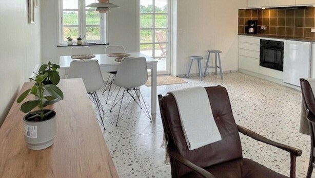 A modern kitchen with white cabinets, a dining area with a wooden table and chairs, and a view of the outdoors through large windows.