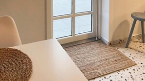 A room with a white table, a woven rug, a grey stool, and a sliding glass door leading to an outdoor area with greenery.