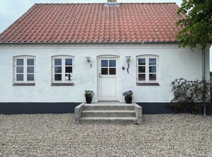 Un bâtiment blanc avec un toit recouvert de tuiles rouges, une porte blanche et deux plantes en pot placées sur les marches de l’entrée.