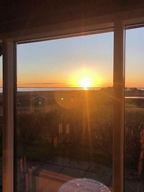 A sunset view from a window overlooking a grassy area with a building in the distance. - Holiday home with sea views over the Western Sea (Vestervig)