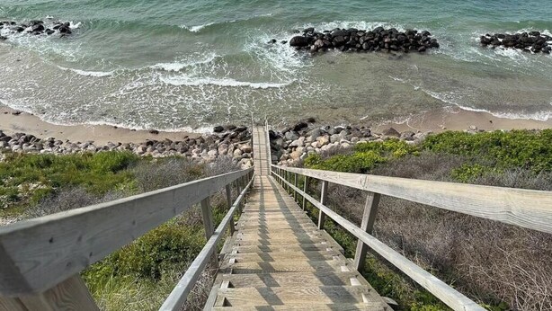 Eine Holztreppe führt zu einem Strand, gesäumt von Felsen und Vegetation, mit einem Geländer.