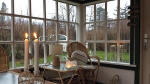 Ein gemütliches Zimmer mit Korbmöbeln, einem Tisch mit Kerzen und Blick ins Freie durch große Fenster.