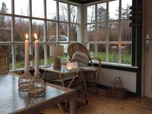 Ein gemütliches Zimmer mit Korbmöbeln, einem Tisch mit Kerzen und Blick ins Freie durch große Fenster.