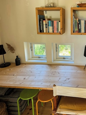 Ein Zimmer mit einem Holztisch, zwei Fenstern, einem Bücherregal, einer Lampe und einer Pflanze.