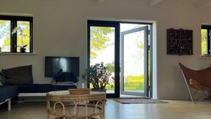 Uma sala de estar com sofá, televisão, mesa de vime e porta de vidro corrediça que dá para um jardim.