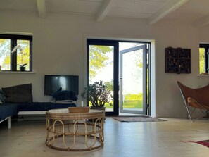 Ein Wohnzimmer mit einer Couch, einem Fernseher, einem Korbstuhl und einer Glastür, die in einen Garten führt.