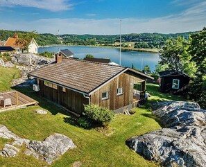 A wooden cabin with a sloped roof, situated on a grassy area with large rocks and a lake in the background. - Cozy cabin with fjord view and fireplace (Uddevalla)