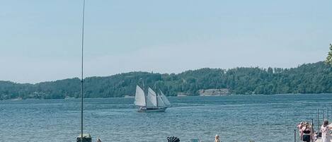 A lakeside view with a sailboat, people relaxing on the shore, and a kite flying.