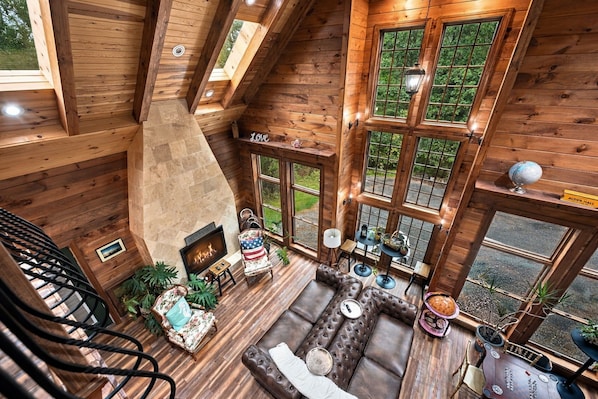 Interior de madera con techos altos, chimenea, zona de estar y grandes ventanales con vistas al verdor.
