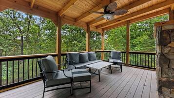 A covered outdoor deck with a wooden ceiling, a stone wall, and a view of a forest.
