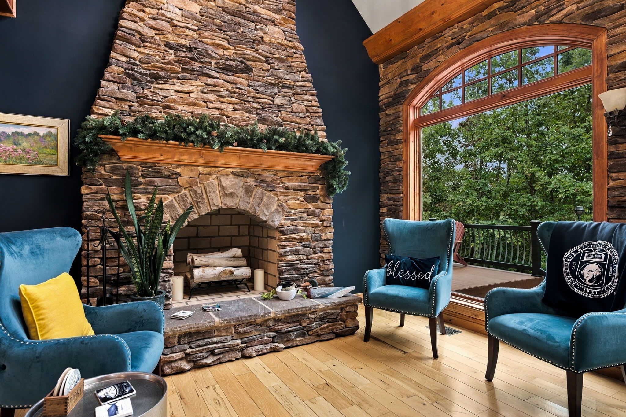 A cozy living room with a stone fireplace, two blue armchairs, and a large window overlooking greenery.