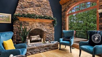 A cozy living room with a stone fireplace, two blue armchairs, and a large window overlooking greenery.