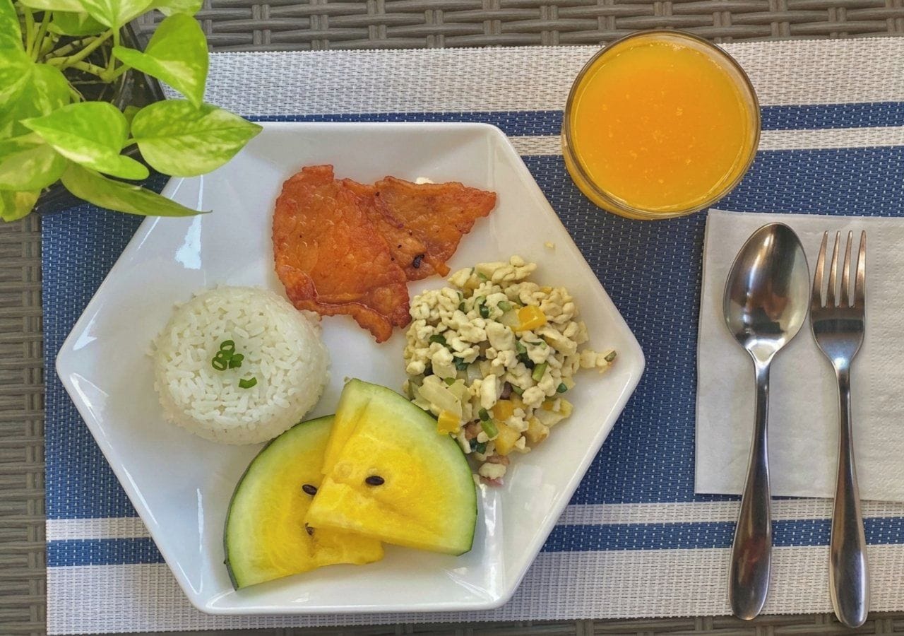 A square plate with white rice, fried bacon, scrambled eggs, and sliced pineapple on a blue and white striped tablecloth.