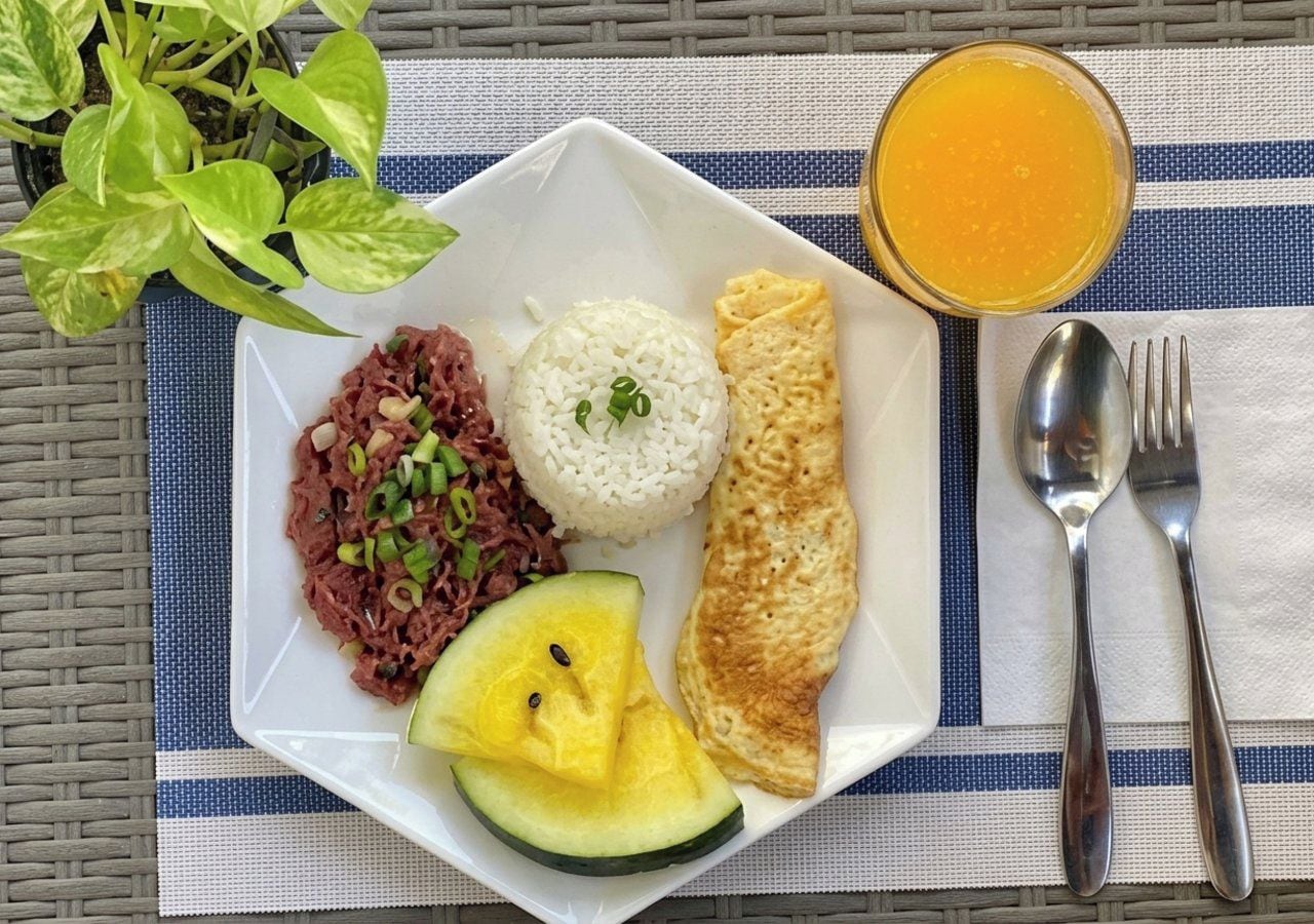 Un repas composé de riz, de viande, d’une omelette et d’ananas, servi dans une assiette, accompagné d’un verre de jus d’orange et de couverts sur une table.