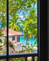 A view through a window of a house with a red roof, surrounded by lush greenery and palm trees, overlooking a clear blue sea.