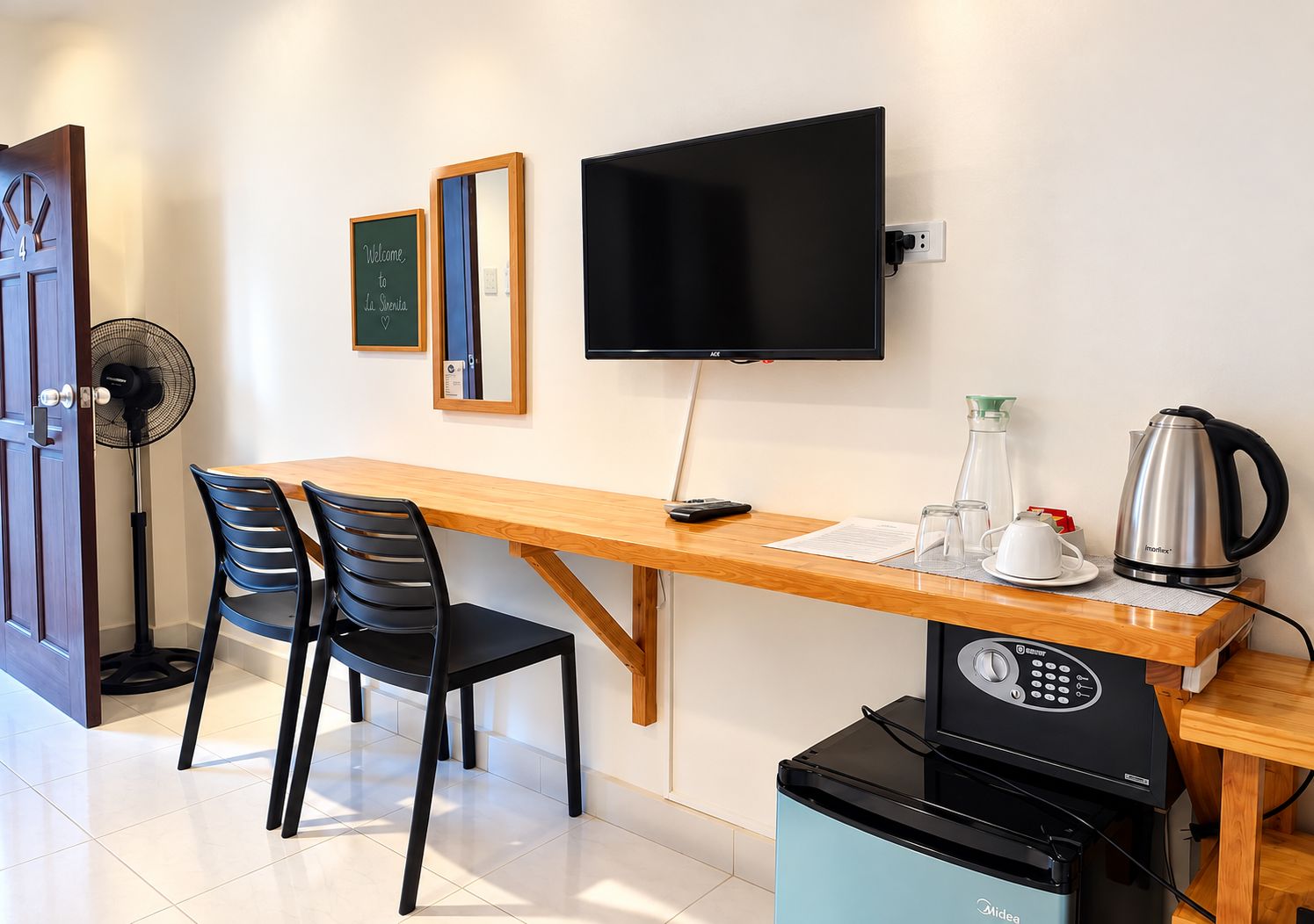 Une pièce comprenant un bureau en bois, deux chaises noires, un téléviseur à écran plat, une bouilloire, une machine à café et un ventilateur.