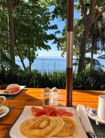 A table with breakfast items, a view of the sea, and tropical vegetation.