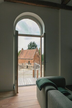 A room with a wooden floor, a sofa, and a large arched window leading to an outdoor area with a brick building. - Attic flat with balcony and sauna in Naulitz (Werben)