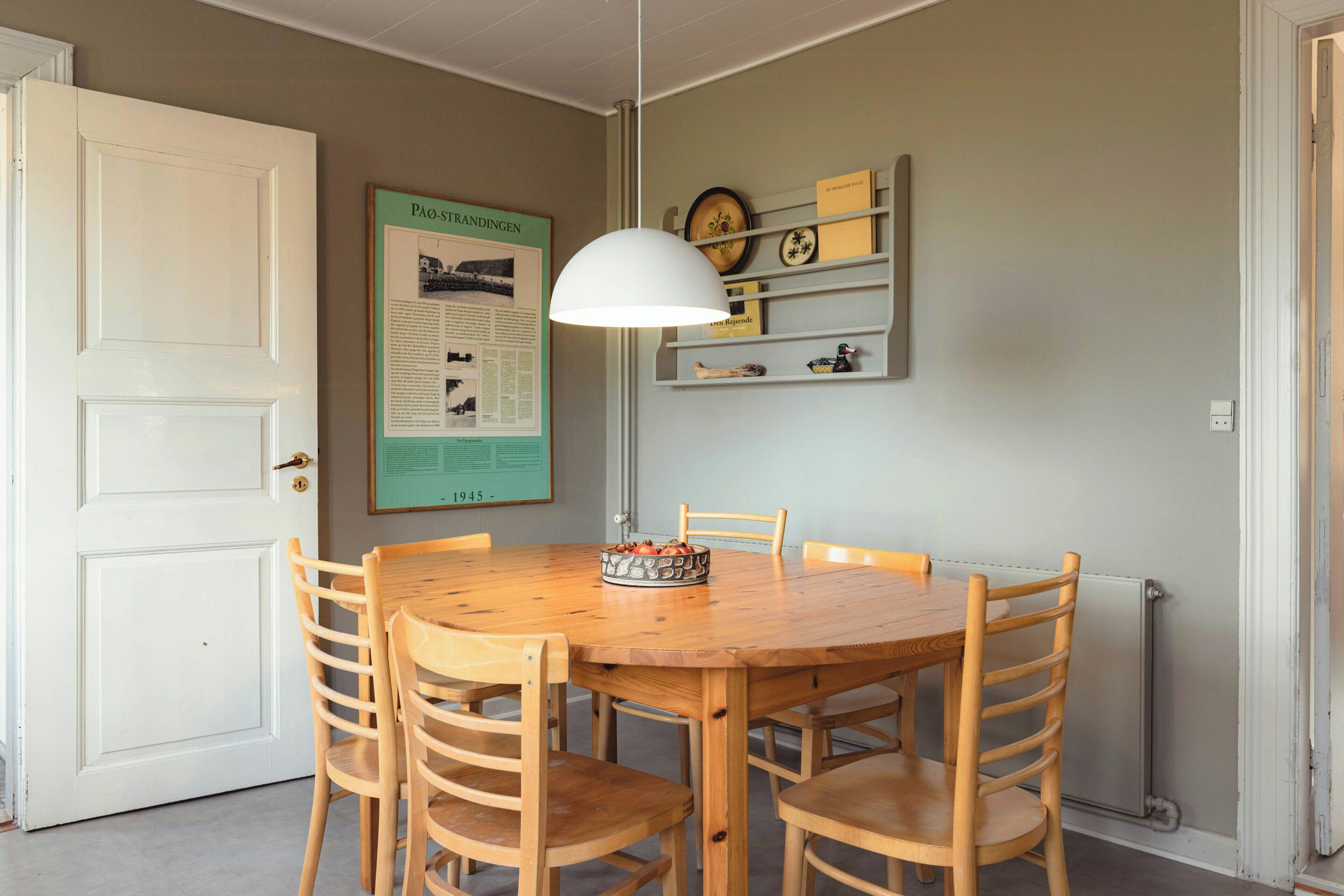 Comedor con mesa y sillas de madera, lámpara colgante, un tablón de anuncios con un cartel enmarcado y un radiador.