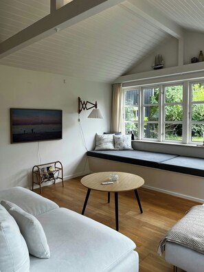 Ein Zimmer mit einer großen Fensterbank, einem runden Holztisch, einem weißen Sofa und einer Wandlampe.