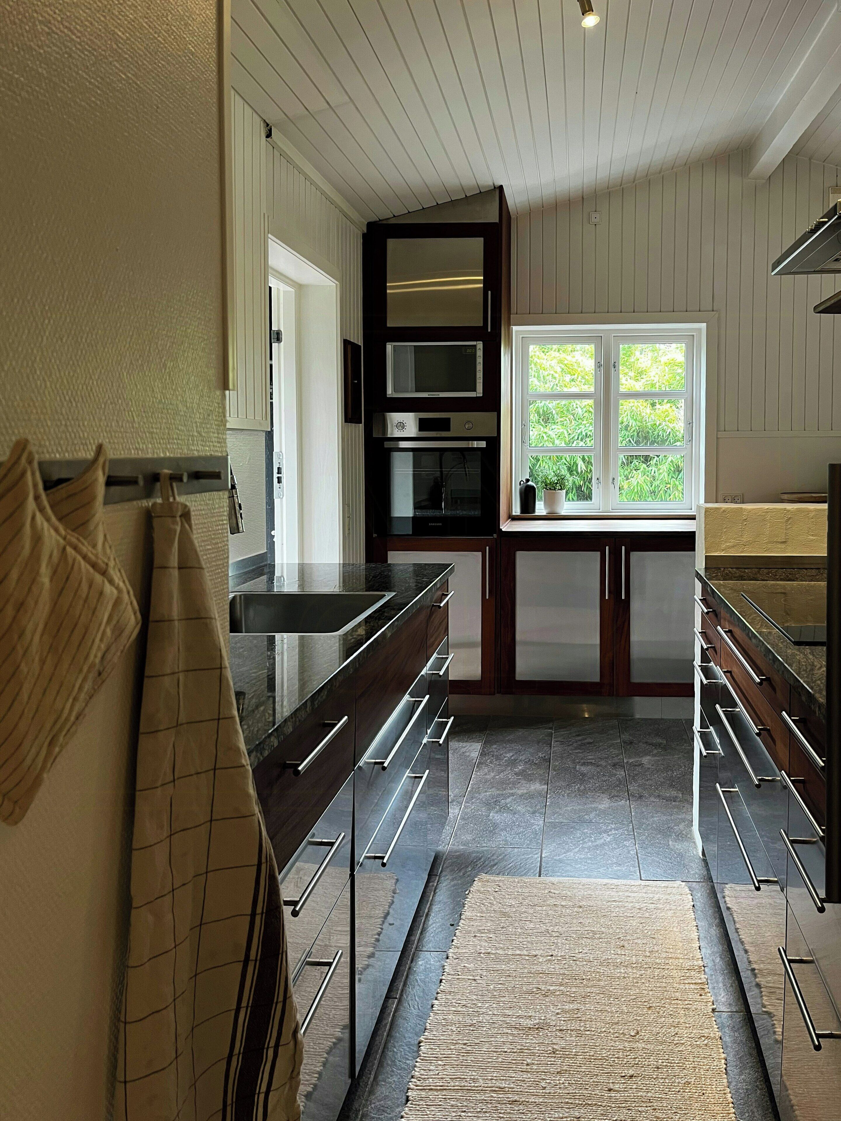 A modern kitchen with built-in appliances, a window with a view of greenery, and a patterned rug on the floor.