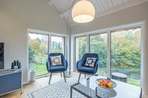 Ein modernes Zimmer mit zwei blauen Stühlen, einem Tisch mit Obst und großen Fenstern mit Blick auf Bäume.