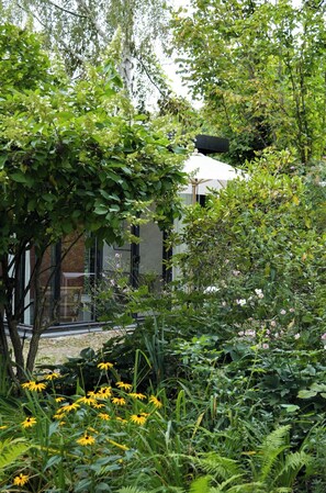 Ein Garten mit verschiedenen Pflanzen und Blumen, ein Baum und ein Gebäude mit weißem Dach.