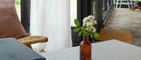 A room with a table, a book, a vase with flowers, a wicker chair, and a view through large windows.