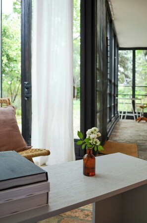 Ein Zimmer mit Tisch, Buch, Blumen in einer Vase, einem Korbstuhl und Blick durch große Fenster.