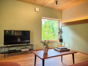 A living room with a TV, a wooden table, a window with a view of greenery, and a potted plant. - Franconian farm house with sauna in Haßberge (Hofheim i. Ufr.)