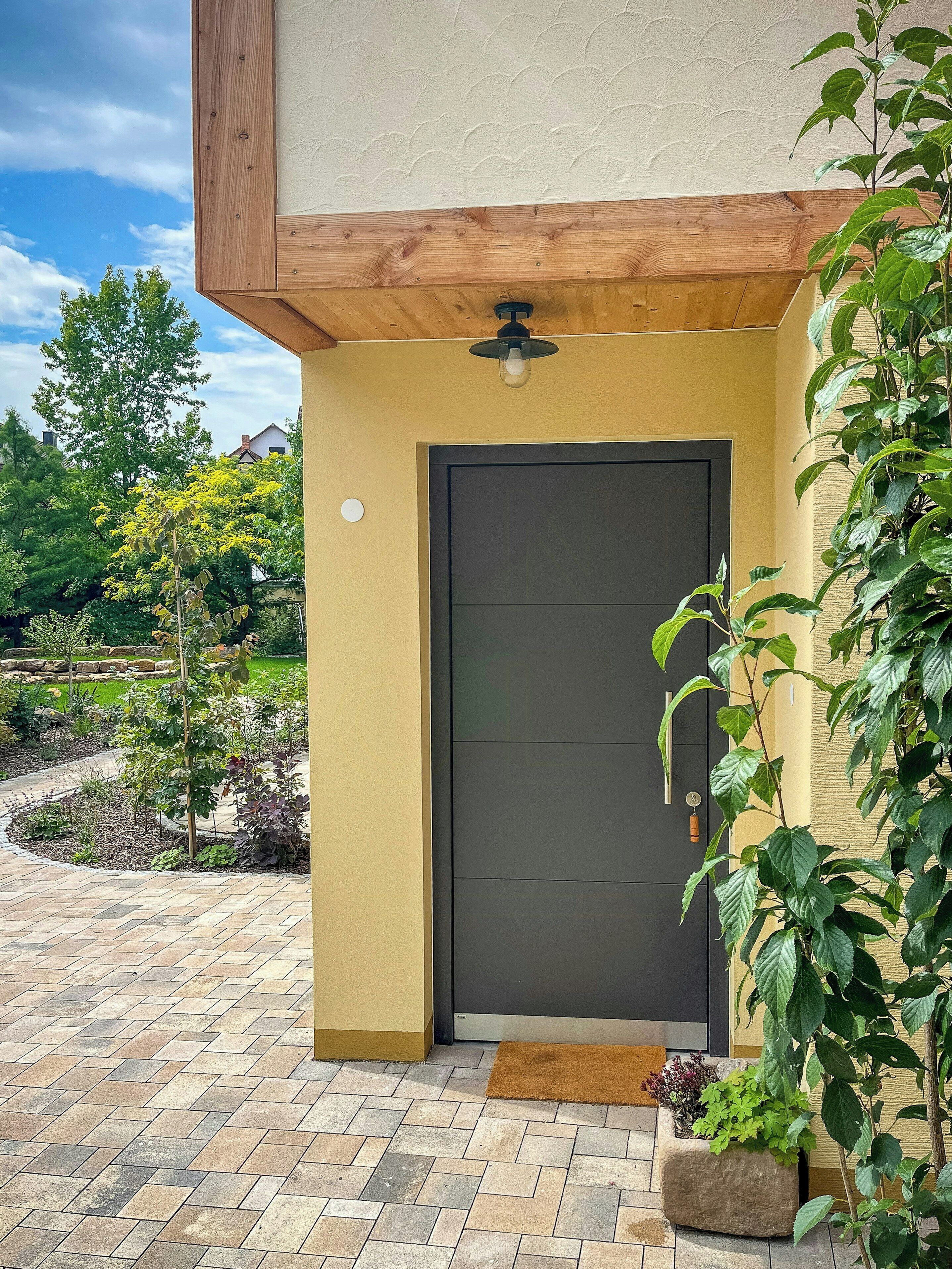 Uma entrada moderna de edifício com porta escura, um pequeno alpendre e um jardim com diversas plantas.