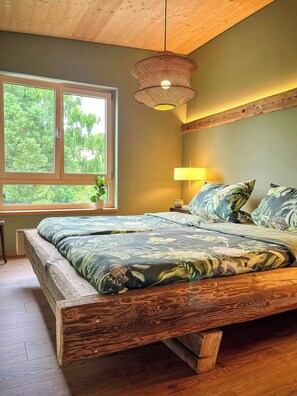 A bedroom with a wooden bed frame, a large window with a view of greenery, and a hanging light fixture. - Franconian farm house with sauna in Haßberge (Hofheim i. Ufr.)
