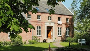 Ein Backsteinhaus mit einem zentralen Eingang, ein kleiner Garten mit einer Bank und ein Weg, der zum Eingang führt.
