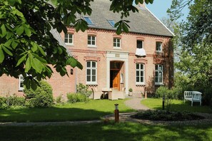 Ein Backsteinhaus mit einem zentralen Eingang, ein kleiner Garten mit einer Bank und ein Weg, der zum Eingang führt.